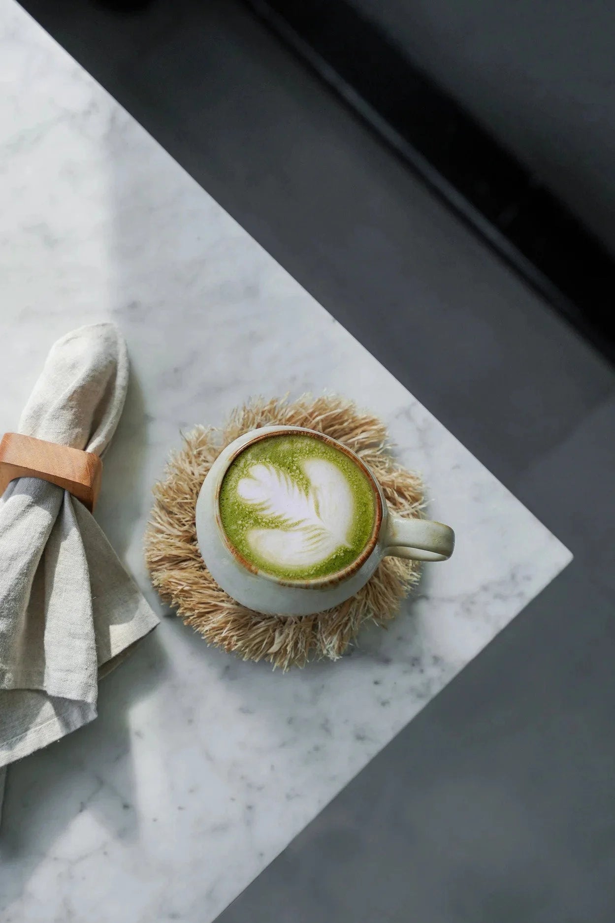 Tasse en céramique blanche pour matcha latte avec motif feuille de thé vert sur mousse, sur dessous de verre rond en raphia naturel à franges solaires
