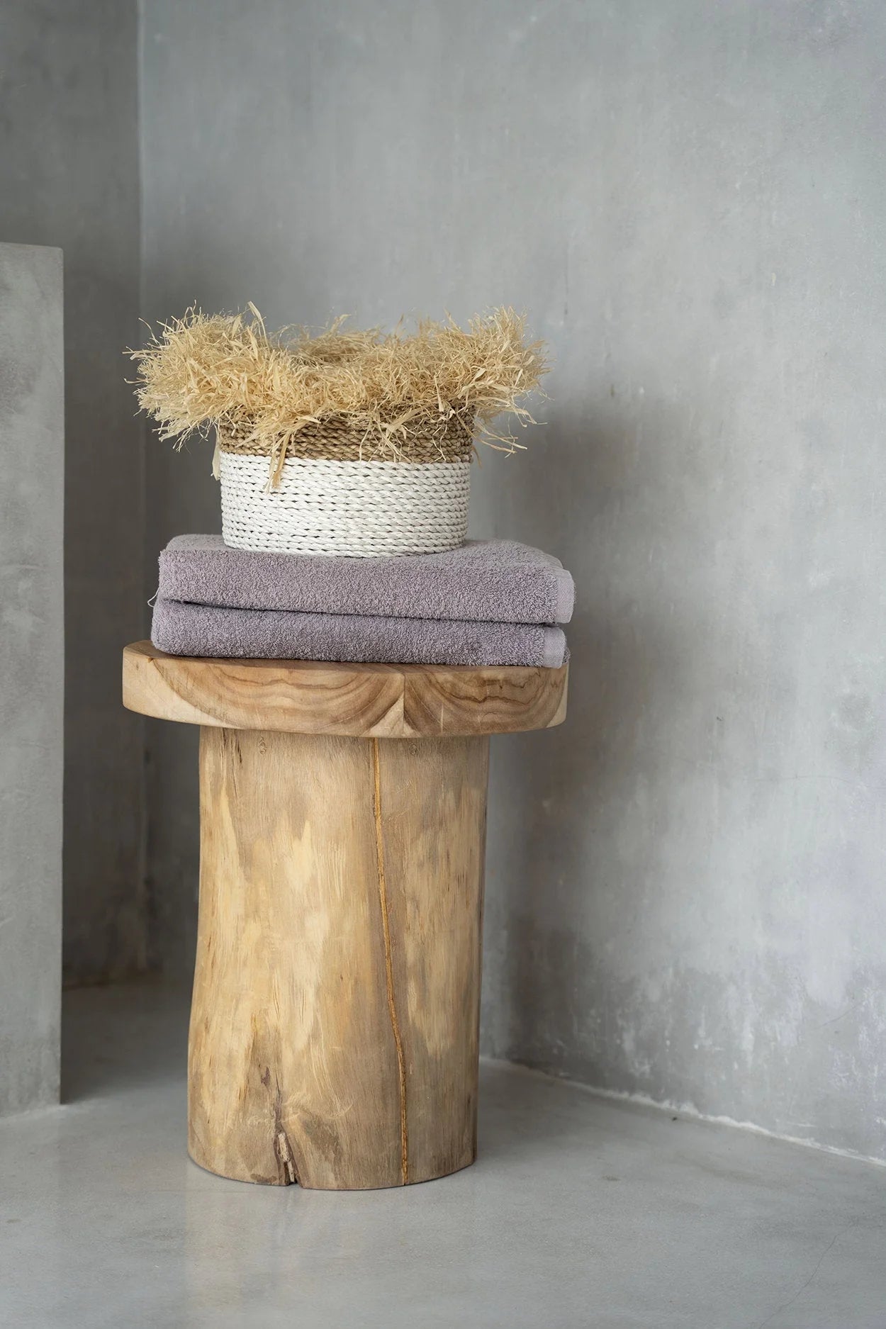 Panier tissé blanc rempli de paille dorée sur pile de serviettes lavande, sur piédestal en bois rustique